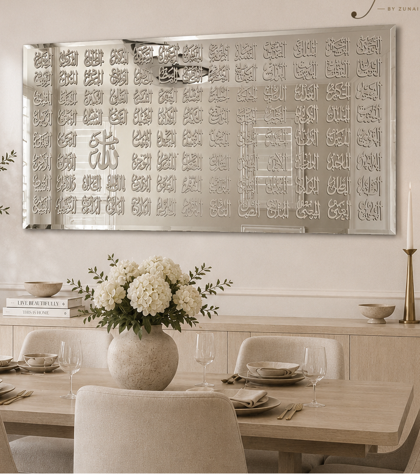 A large rectangular wall mirror with intricate Arabic calligraphy patterns around the border hangs on a white wall in a styled dining room. The room features a light wood dining table, neutral upholstered chairs, a vase of white flowers, and other tasteful home decor accessories.