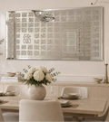A large rectangular wall mirror with intricate Arabic calligraphy patterns around the border hangs on a white wall in a styled dining room. The room features a light wood dining table, neutral upholstered chairs, a vase of white flowers, and other tasteful home decor accessories.