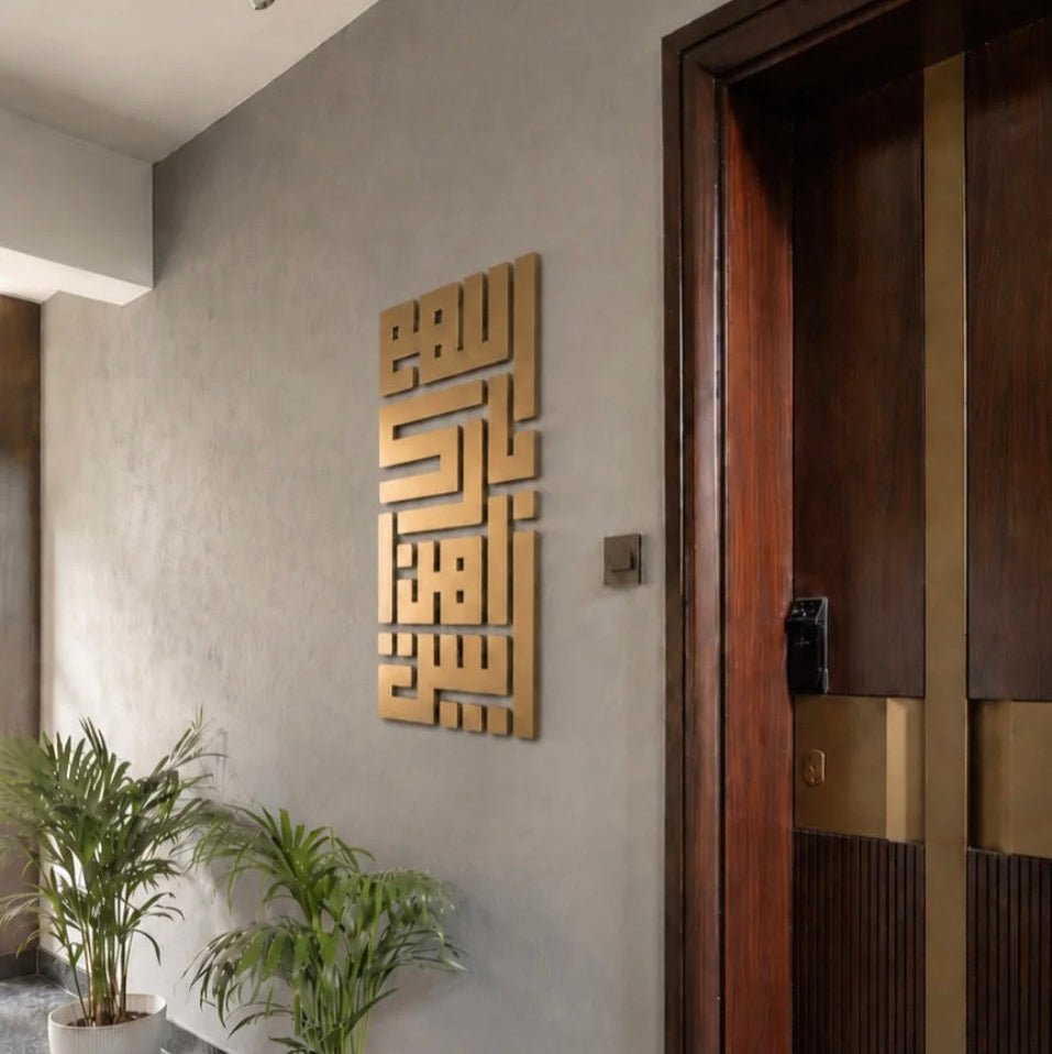 A vertical gold-toned geometric wall art piece mounted on a textured gray wall in a modern interior hallway, with potted plants and a dark wood door visible.
