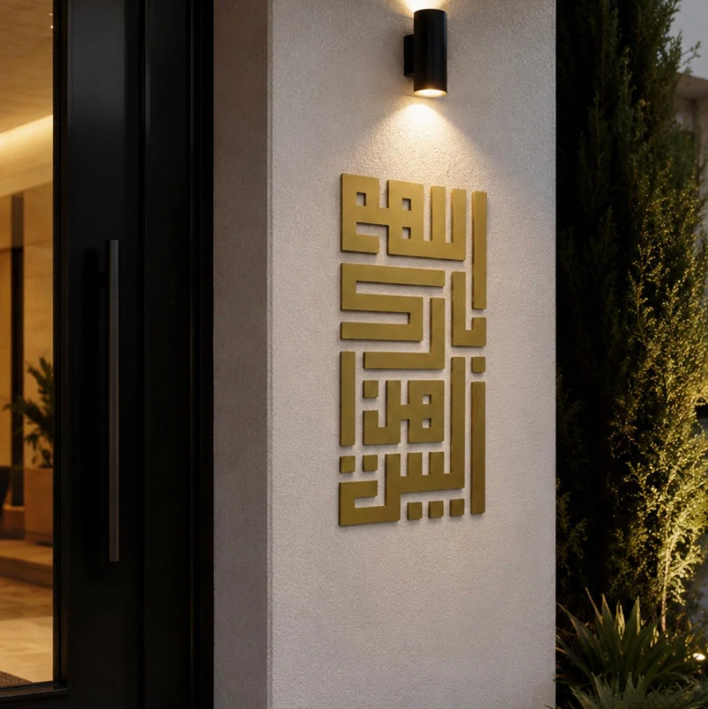 A modern black wall sconce mounted on a textured beige wall above gold Arabic calligraphy, next to a black glass door and greenery at night.