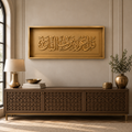 A rectangular gold-toned wall art piece with raised Arabic calligraphy Qul Auzu bi rabbil falaq hangs above a carved wooden console table in a styled interior room. A lamp, books, a bowl, and a vase with branches sit on the table against a beige wall.