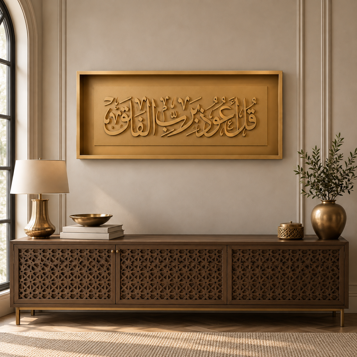 A rectangular gold-toned wall art piece with raised Arabic calligraphy Qul Auzu bi rabbil falaq hangs above a carved wooden console table in a styled interior room. A lamp, books, a bowl, and a vase with branches sit on the table against a beige wall.