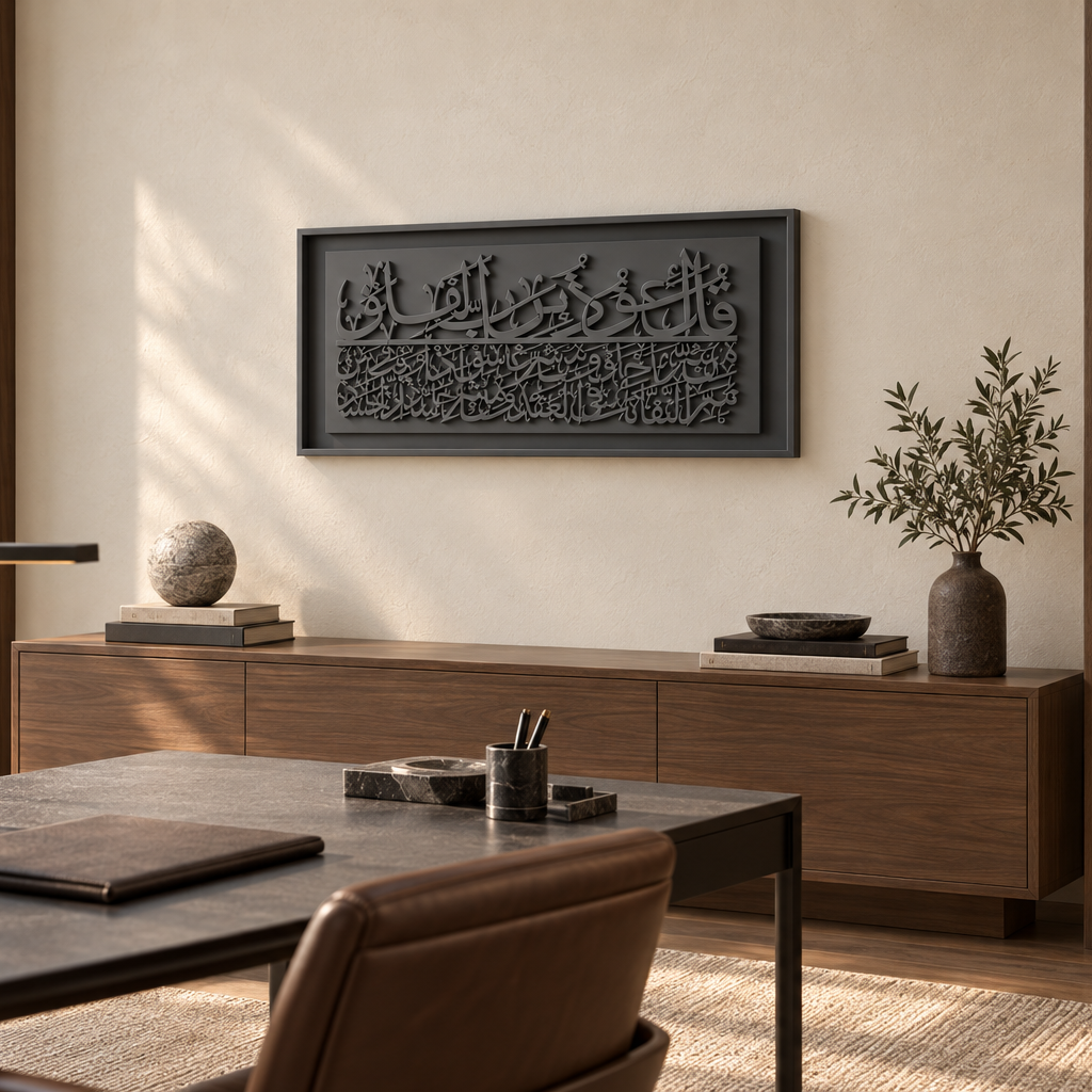 A framed black wall art piece with Arabic calligraphy hangs on a beige wall in a modern, sunlit home office. The room features a wooden credenza, a desk with a leather chair, and a built-in bookshelf with warm lighting.