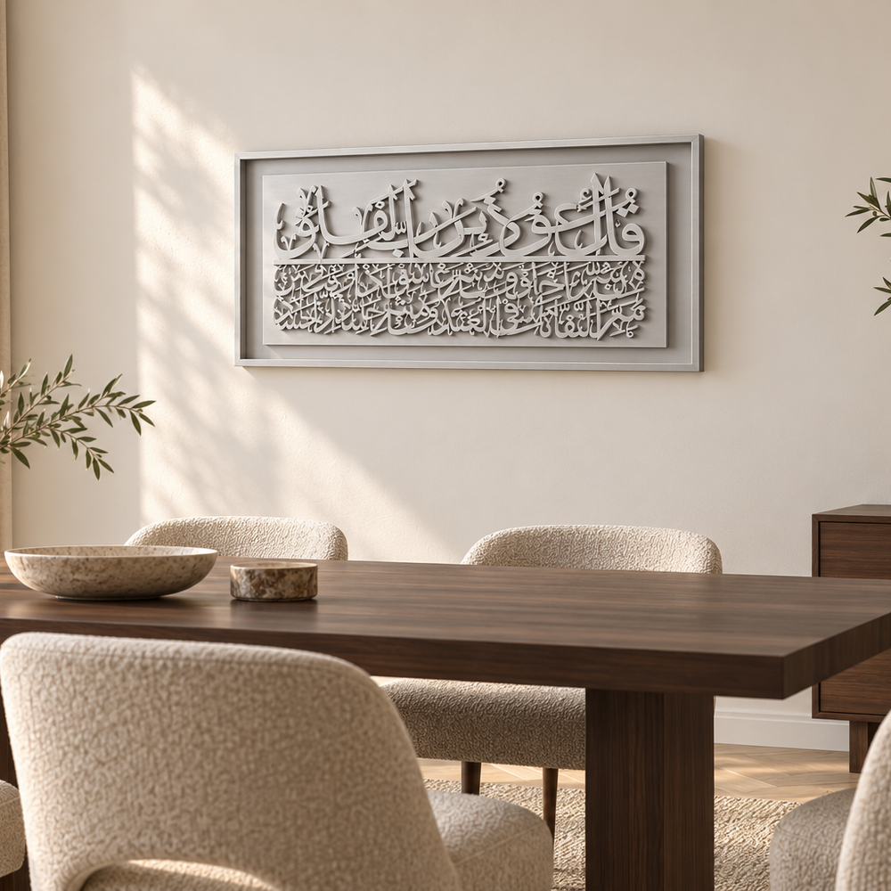 A framed Arabic calligraphy wall art piece in a light gray tone, displayed in a modern dining room setting above a dark wood dining table and textured beige chairs.