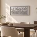 A framed Arabic calligraphy wall art piece in a light gray tone, displayed in a modern dining room setting above a dark wood dining table and textured beige chairs.