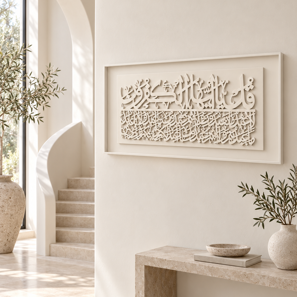 A rectangular framed Islamic calligraphy wall art piece in a warm cream tone, displayed on a beige stone bench in a bright, modern minimalist entryway with a staircase and archway in the background.