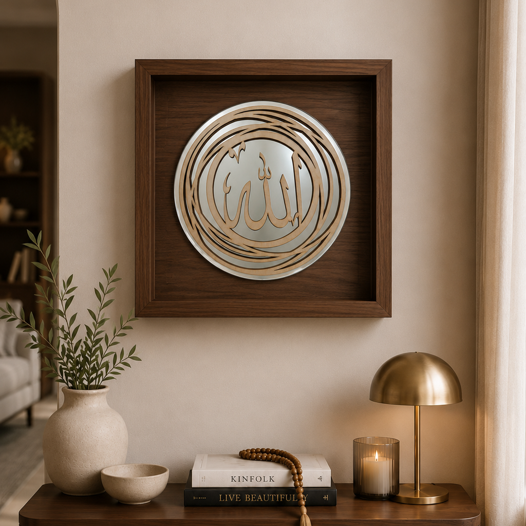 A square framed Islamic wall art piece featuring gold Arabic calligraphy on a silver circular background, mounted on a beige wall above a wooden console table styled with a vase, books, a candle, and a brass table lamp.