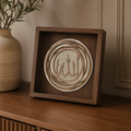 A square wooden shadow box display featuring layered circular wood cutouts with Arabic calligraphy, placed on a wooden cabinet next to a beige ceramic vase with green branches.