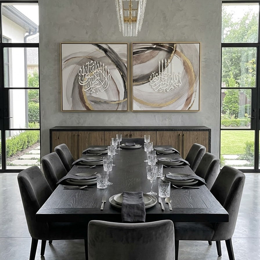 A modern dining room featuring a dark wood rectangular table set with silverware and glassware, surrounded by dark gray velvet chairs. Above the table, two large framed abstract paintings with Arabic calligraphy hang on a textured gray wall, flanked by tall black-framed windows.