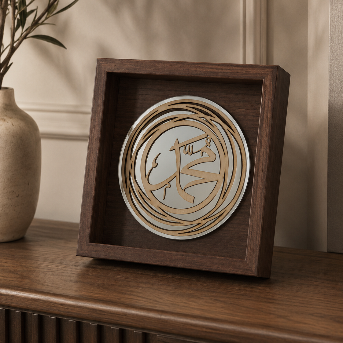 A square dark wood shadow box display featuring a circular layered wooden design with Arabic calligraphy, shown on a wooden console table next to a ceramic vase.